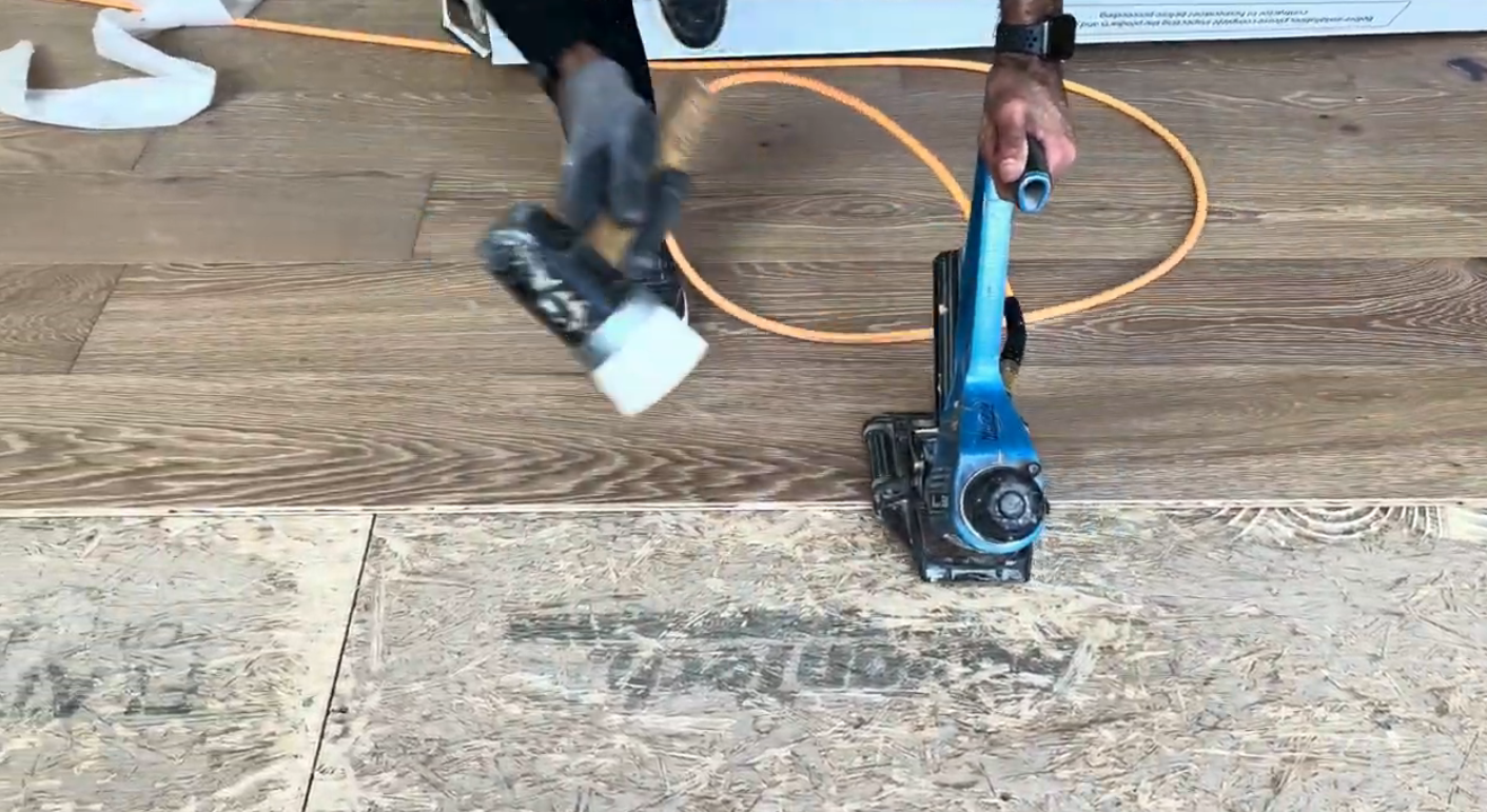 Person using a blue flooring nailer and a mallet on engineered hardwood flooring, with visible wood grain and installation materials in a home setting.