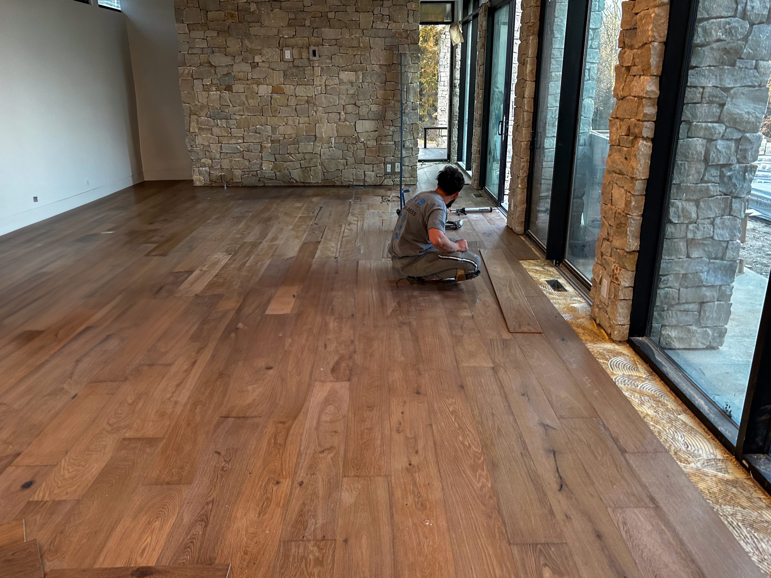 Man installing hardwood flooring in a modern room with stone walls and large windows, showcasing the Palms Collection by SVB Wood Floors.