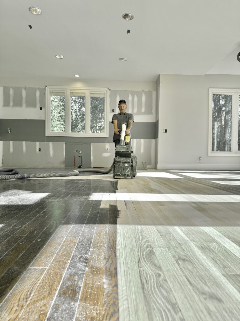Man operating a dustless hardwood floor refinishing machine in a partially renovated room, showcasing contrasting sections of refinished wood floors.