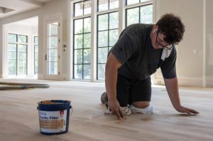 Contractor repairing dents in hardwood floor with wood filler and tools.